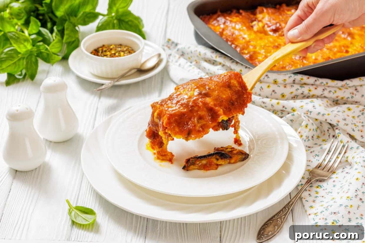25 Comfort-Food Recipes That Satisfy Every Craving 15 A hand serves a slice of Eggplant Parmigiana onto a white plate, with a casserole dish, herbs, and condiments visible on a white table.