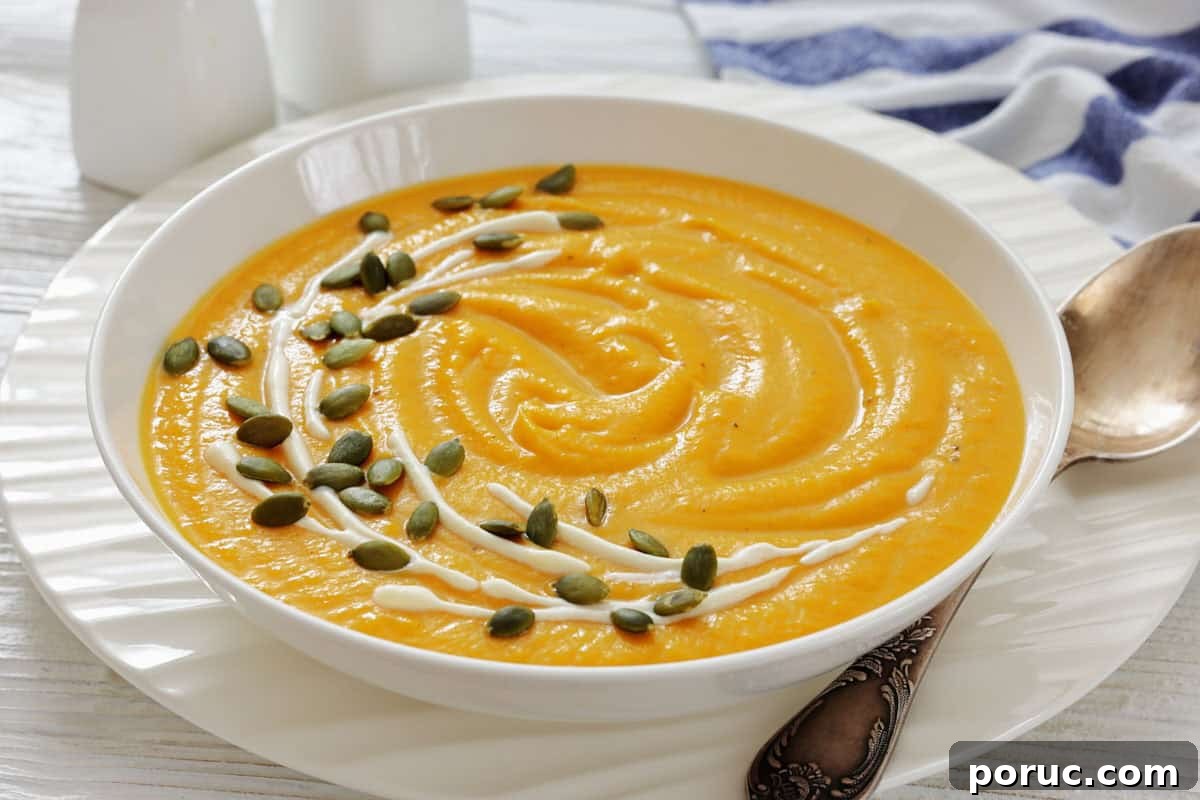 25 Comfort-Food Recipes That Satisfy Every Craving 25 A bowl of creamy pumpkin soup with canned pumpkin garnished with a swirl of cream and sprinkled with pumpkin seeds, placed on a white plate beside a spoon.