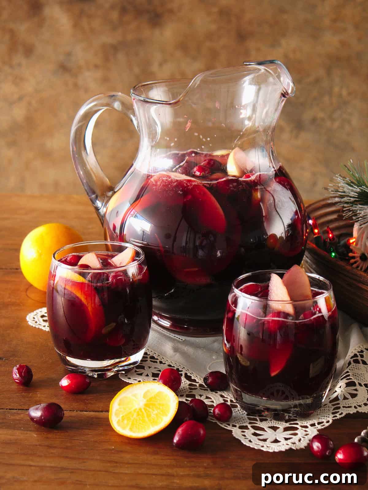 Clear glass pitcher and two rocks glasses filled with fruit-filled red Christmas sangria on a wooden table, styled with scattered cranberries, a white doily, and a bowl of multicolored Christmas lights.