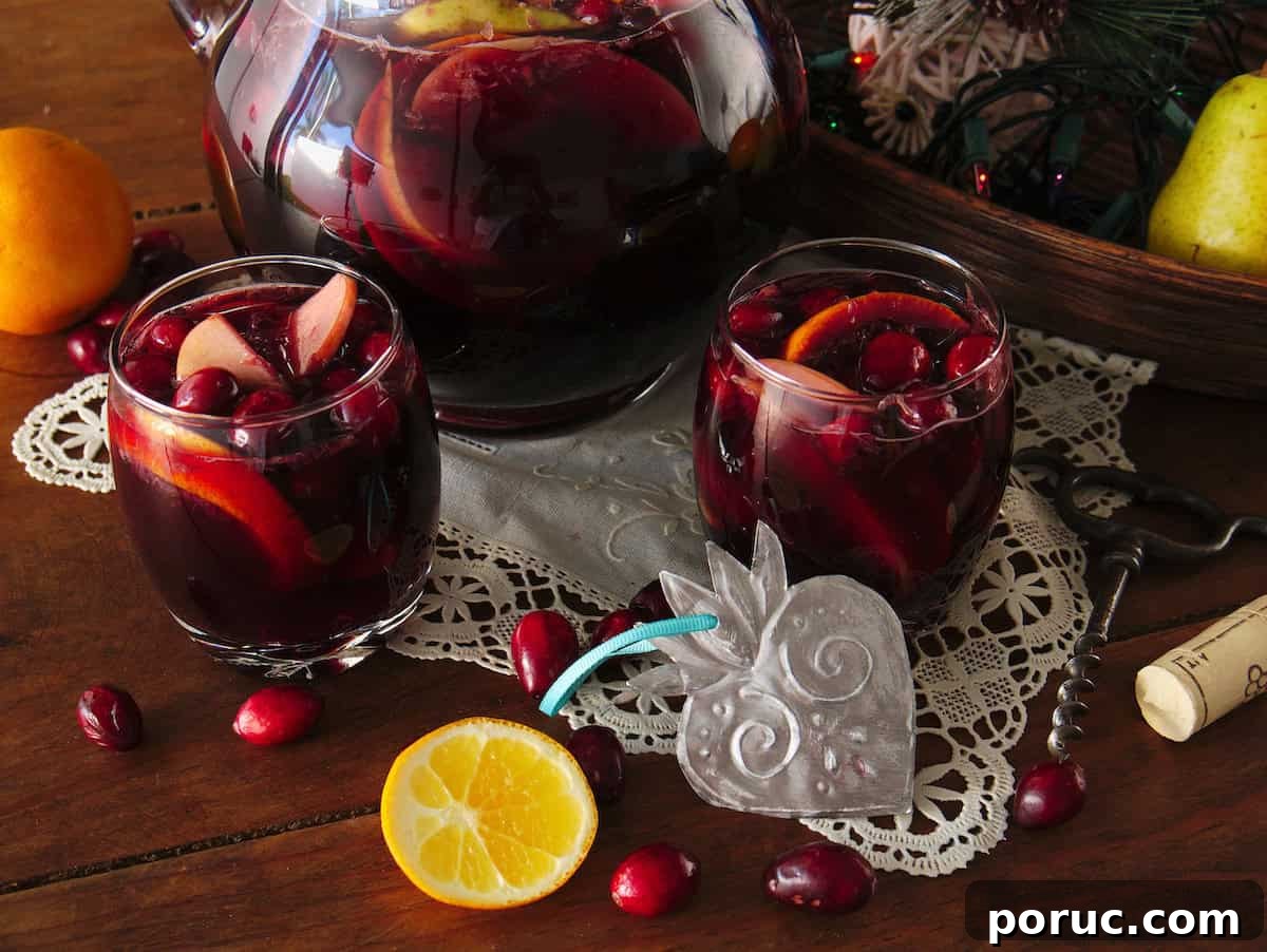 Wooden table topped with a white cloth doily, featuring a pitcher and two rocks glasses filled with Christmas sangria, with fruit scattered around.