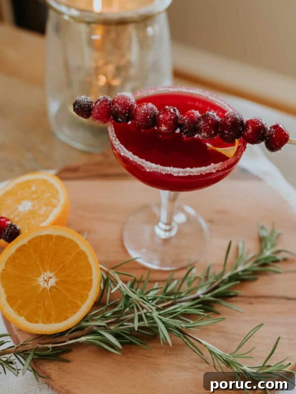 A festive cranberry margarita served with a skewer of sugared cranberries and a sugared rim in a martini class on a wooden cutting board with rosemary and orange slices as garnish.