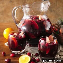 Clear glass pitcher and two rocks glasses filled with fruit-filled red Christmas sangria on a wooden table, styled with scattered cranberries, a white doily, and a bowl of multicolored Christmas lights.