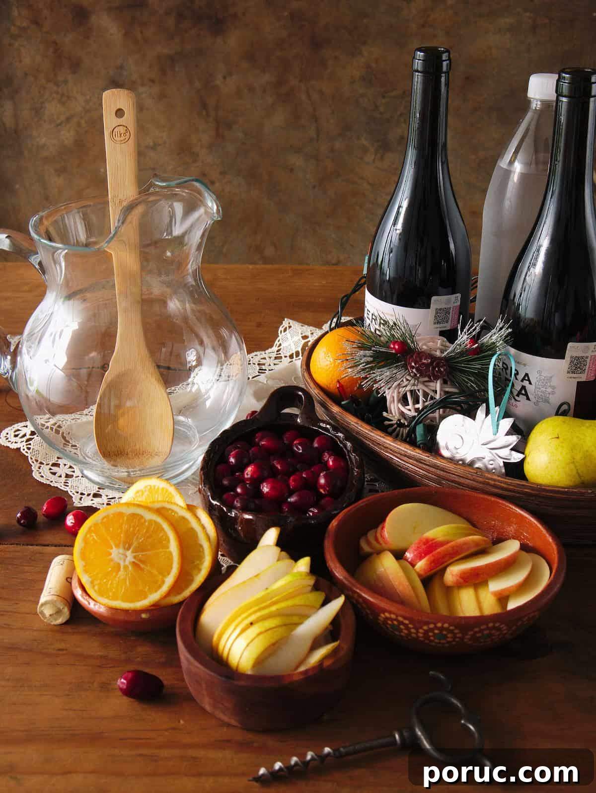 Empty clear glass pitcher with a wooden spoon, bottles of red wine, sliced oranges, pears, and apples, and whole cranberries in earthenware bowls, prepped for making red Christmas sangria.