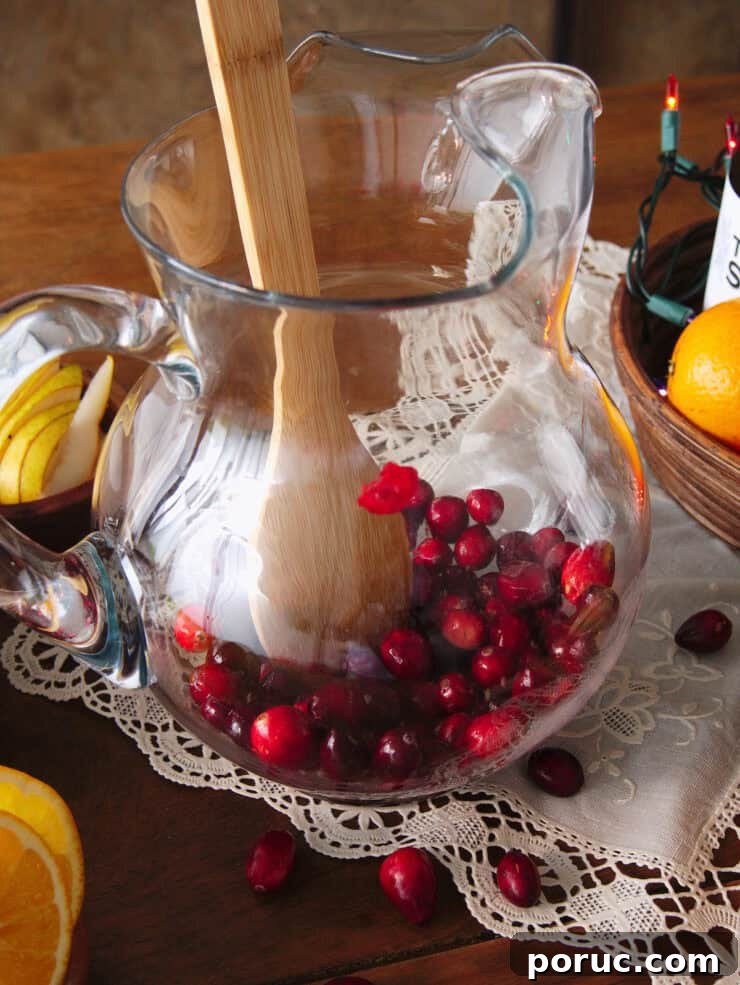Wooden spoon gently muddling fresh cranberries in the bottom of a clear glass pitcher.