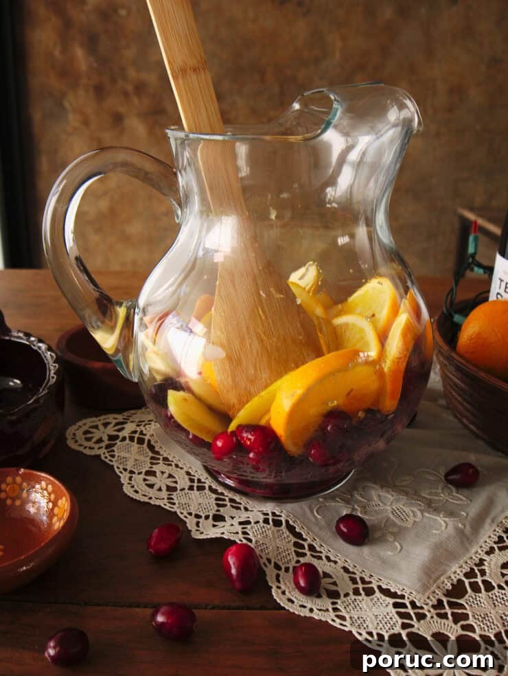 Wooden spoon gently muddling orange slices over fresh cranberries in a clear glass pitcher.