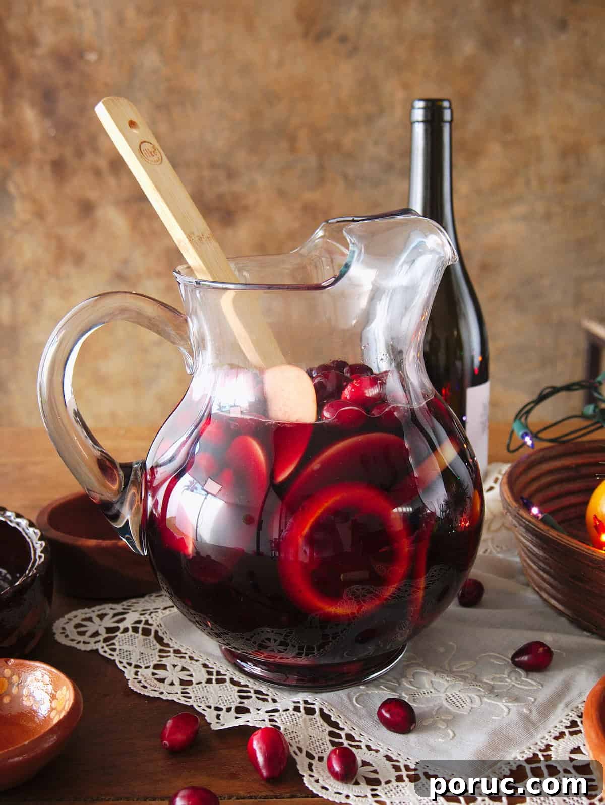 Clear glass pitcher filled about two-thirds with red wine and muddled fruit soaking overnight for Christmas sangria, with a bottle of red wine in the background and fresh cranberries in the foreground.