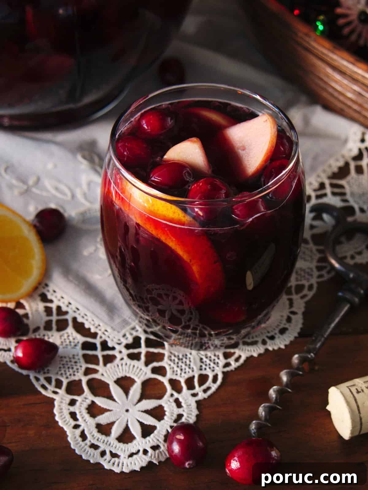 Clear rocks glass filled with red Christmas sangria and fruit, set on a white cloth doily on a wooden table with the sangria pitcher in the background.