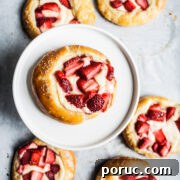 Strawberry Cream Cheese Brioche with Orange Blossom Water 10 Brioche pastries with strawberries and cream cheese on white parchment paper. One pastry is elevated on a white ceramic plate.