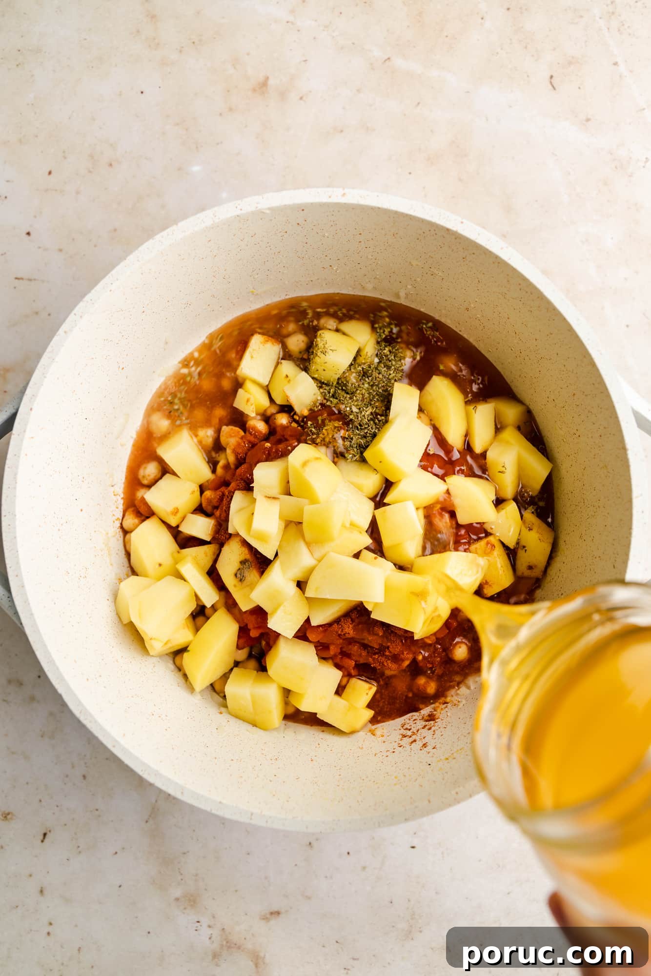Hearty Italian Chickpea Soup Recipe for Comforting Weeknight Meals 6 pouring broth into a white pot full of diced potatoes, herbs, tomatoes, and chickpeas.
