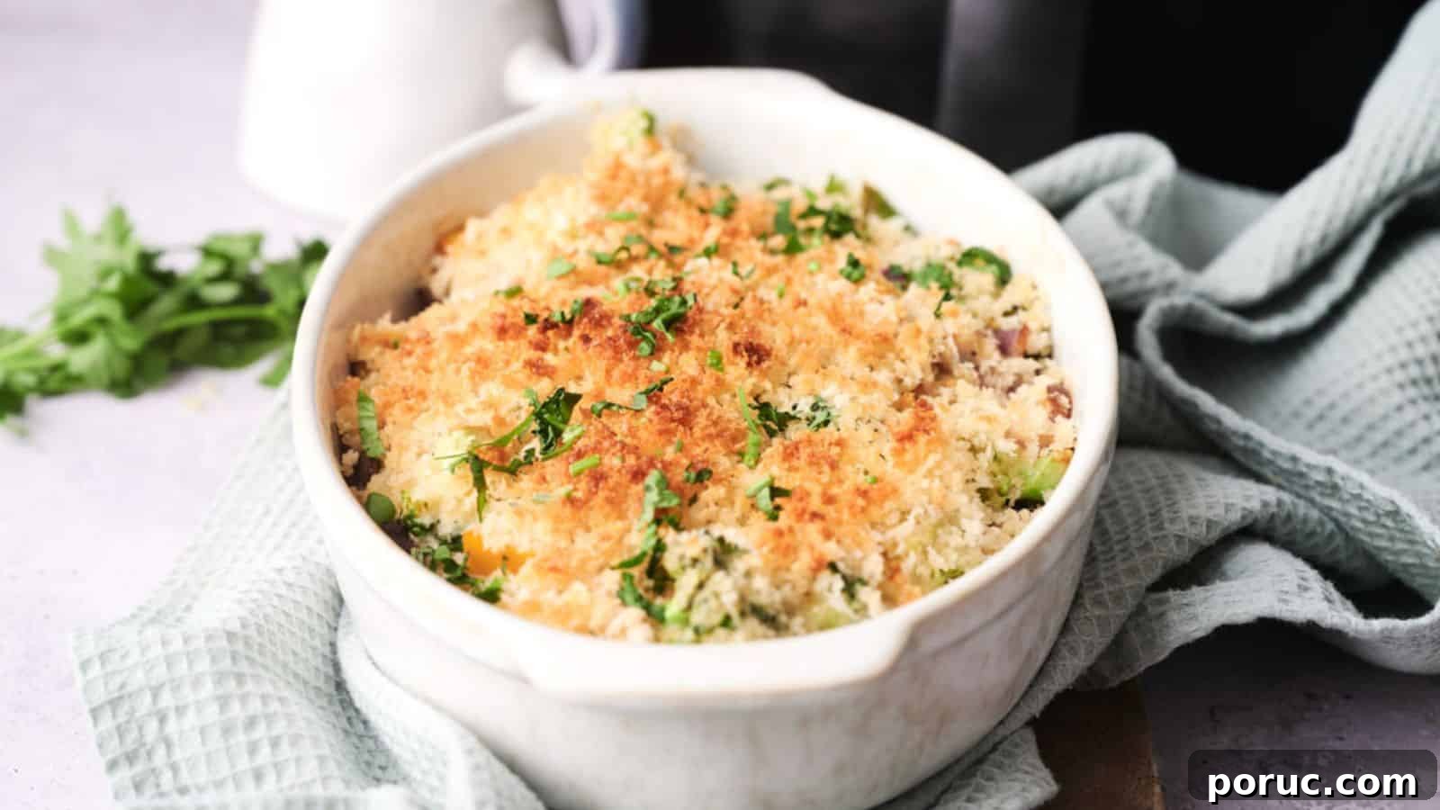 29 Air Fryer Casseroles Perfect for Busy Weeknight Dinners 29 A white ceramic dish containing baked vegetable gratin topped with browned breadcrumbs and parsley, placed on a light green textured cloth.