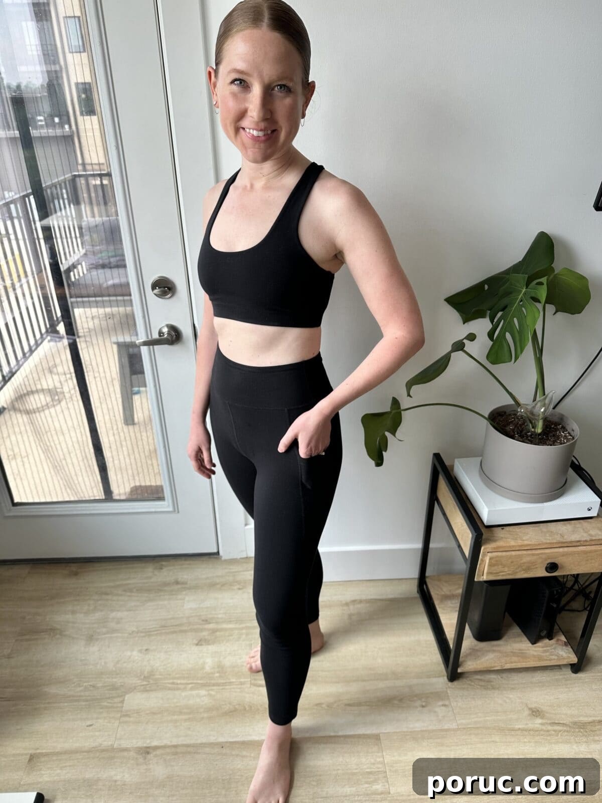 A barefoot woman with her hair pulled back is standing indoors, smiling, and wearing a black sport bra with high-waisted leggings from Mate that have a pocket on the side.