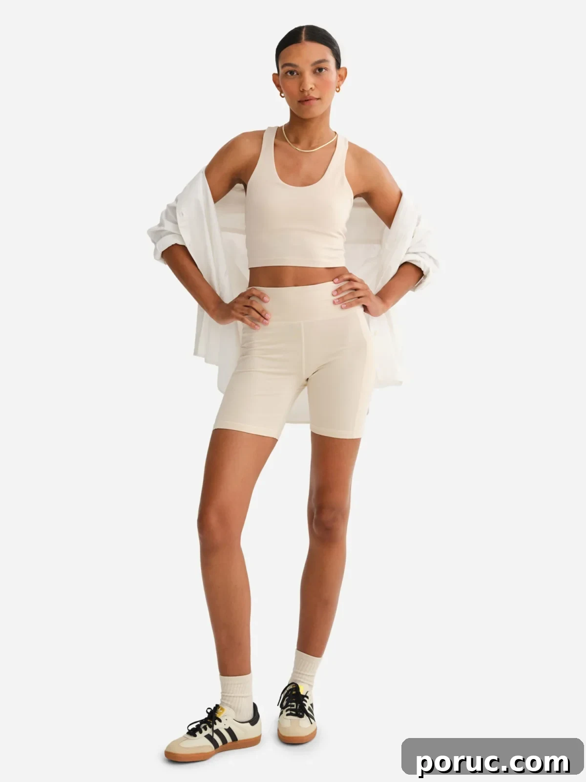 A model with her hands on her hips is wearing a two-piece neutral-toned athleisure set consisting of a cropped tank top and mid-thigh shorts from Mate, paired with an unbuttoned white shirt and sneakers on a white background.