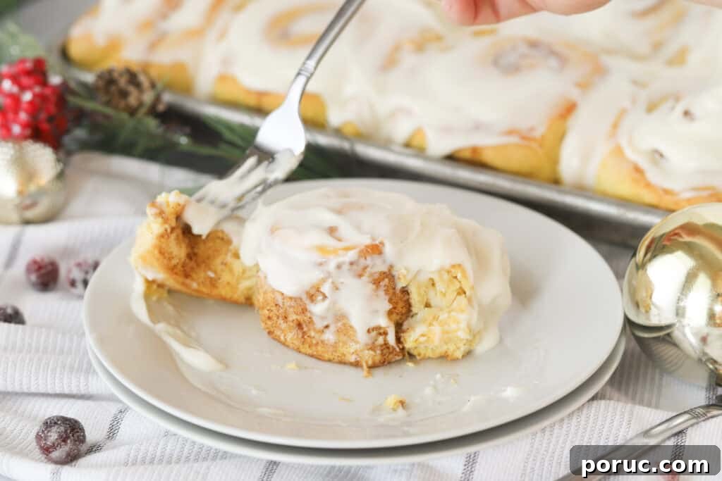 A cinnamon roll on a white plate being eaten with a fork, with a full tray of cinnamon rolls in the background.