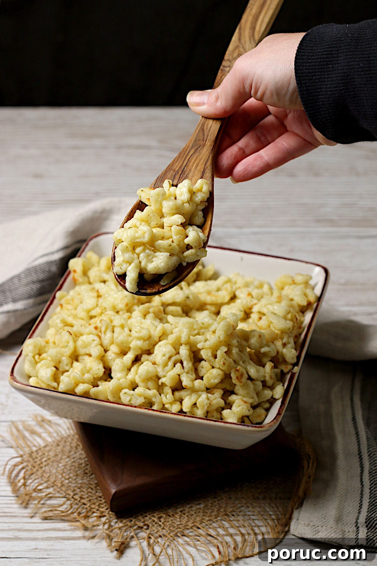 Authentic German Spaetzle Recipe: Step-by-Step Homemade Spaetzle 5 A hand scooping some Spaetzle from a square bowl with a wooden spoon.