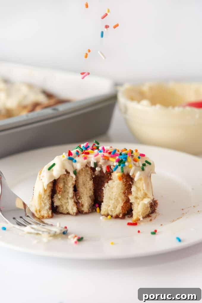 Cream Cheese Frosting Recipe for Perfect Cinnamon Rolls 5 A single cinnamon roll on a white plate, pulled apart to reveal its layers, topped with cream cheese frosting and rainbow sprinkles