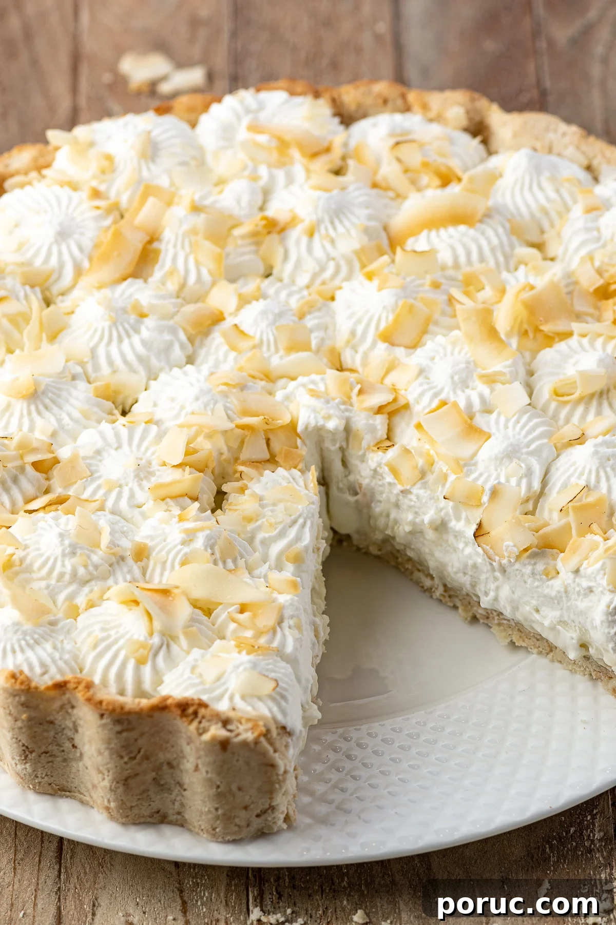 A close up shot of a coconut cream pie on a white texture plate with a slice removed.