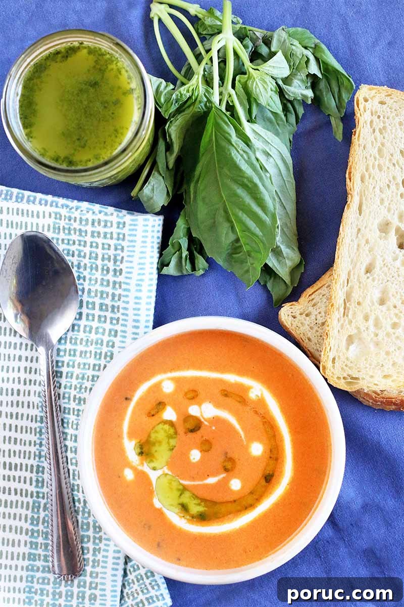 Creamy Tomato Basil Bisque That Elevates Classic Comfort Food 2 Top-down shot of a bowl of tomato bisque garnished with cream and basil oil, with a spoon on a folded gray and white cloth napkin, a jar of green herb oil, sprigs of fresh basil, and slices of sourdough bread, on a blue cloth background.