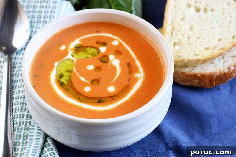 Creamy Tomato Basil Bisque That Elevates Classic Comfort Food 15 A bowl of creamy tomato basil bisque with a cream and herb oil garnish, next to a spoon on top of a folded white and gray cloth napkin, on a blue background with two pieces of bread.