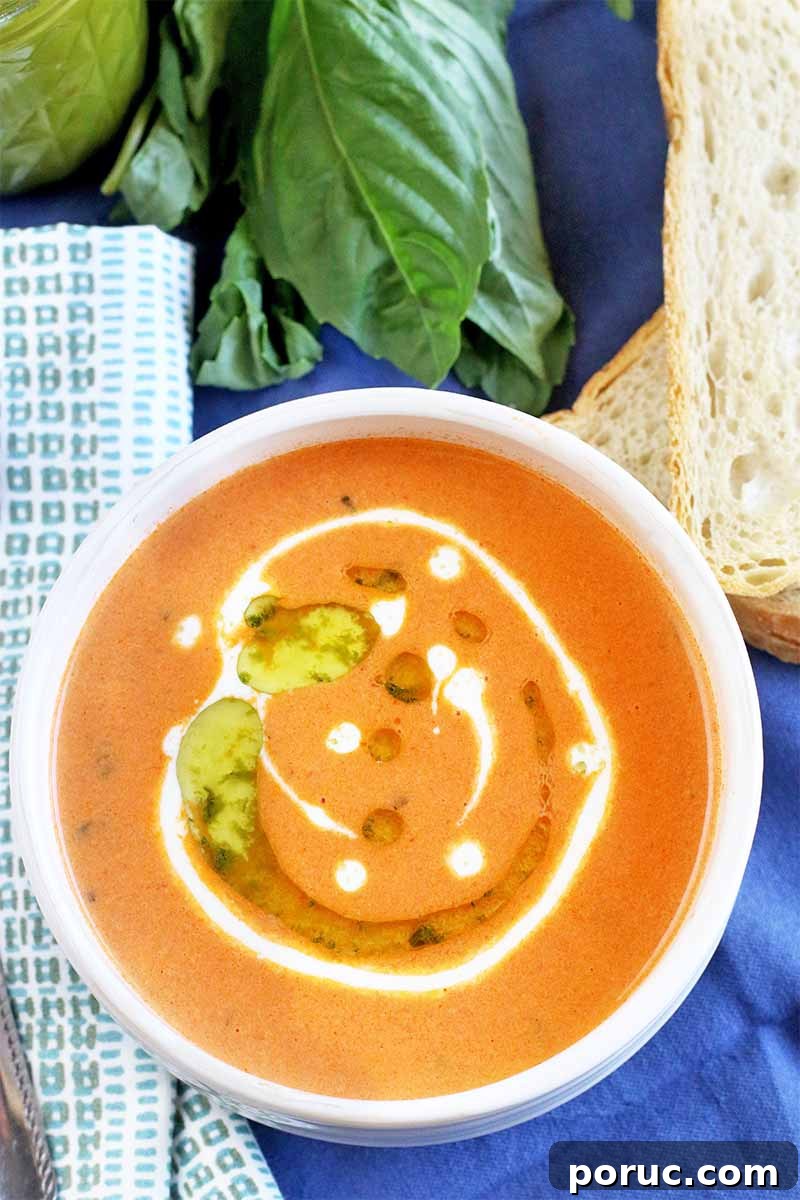 Creamy Tomato Basil Bisque That Elevates Classic Comfort Food 3 Top-down shot of a bowl of tomato bisque garnished with a drizzle of cream and basil oil, on a blue cloth background with a sprig of basil, jar of herb oil, slices of sourdough bread, a folded white and gray cloth napkin, and a spoon.