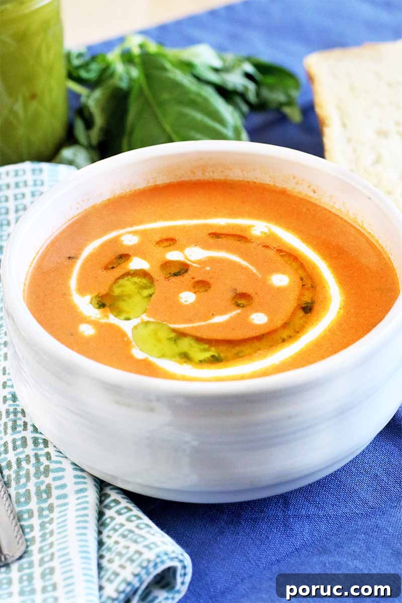 Creamy Tomato Basil Bisque That Elevates Classic Comfort Food 4 Closely cropped shot of a white soup bowl of pale orange tomato bisque, garnished with heavy cream and herb oil, with a jar of more oil and a sprig of basil in the background, with a folded cloth napkin topped with a piece of silverware to the left, on a beige surface topped with a blue cloth.