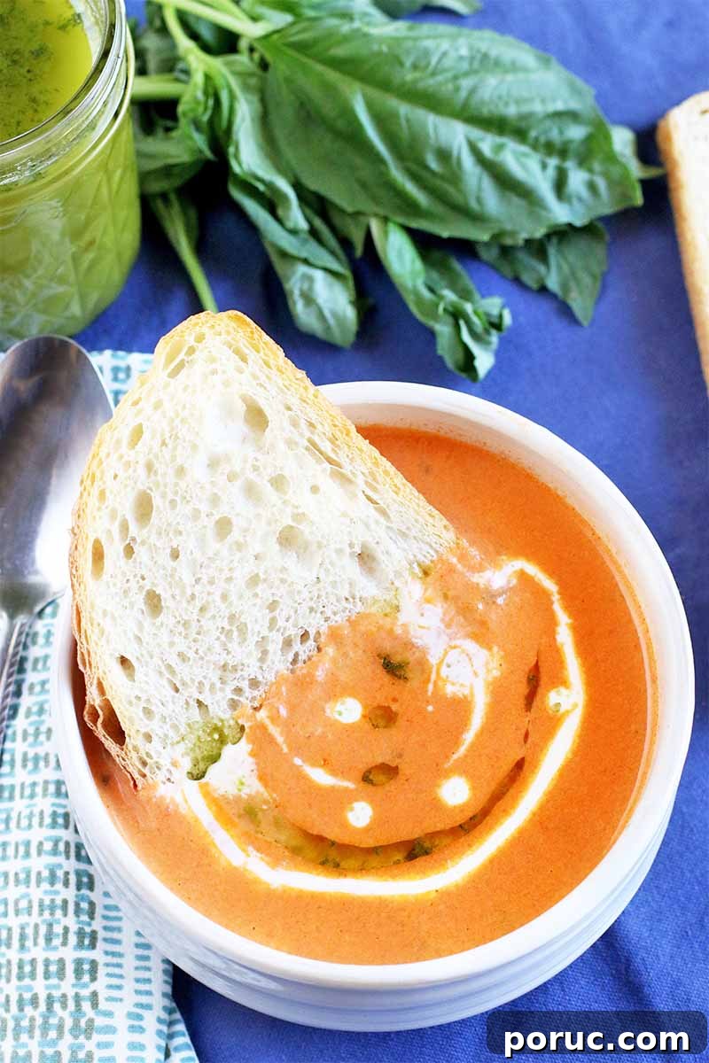 Creamy Tomato Basil Bisque That Elevates Classic Comfort Food 5 A slice of sourdough bread is half submerged in a white ceramic bowl of pale orange bisque garnished with heavy cream and herb oil, with a jam jar of more of the green oil in the background beside a sprig of basil, next to a folded white and gray striped cloth napkin with a spoon on top, on a blue tablecloth.