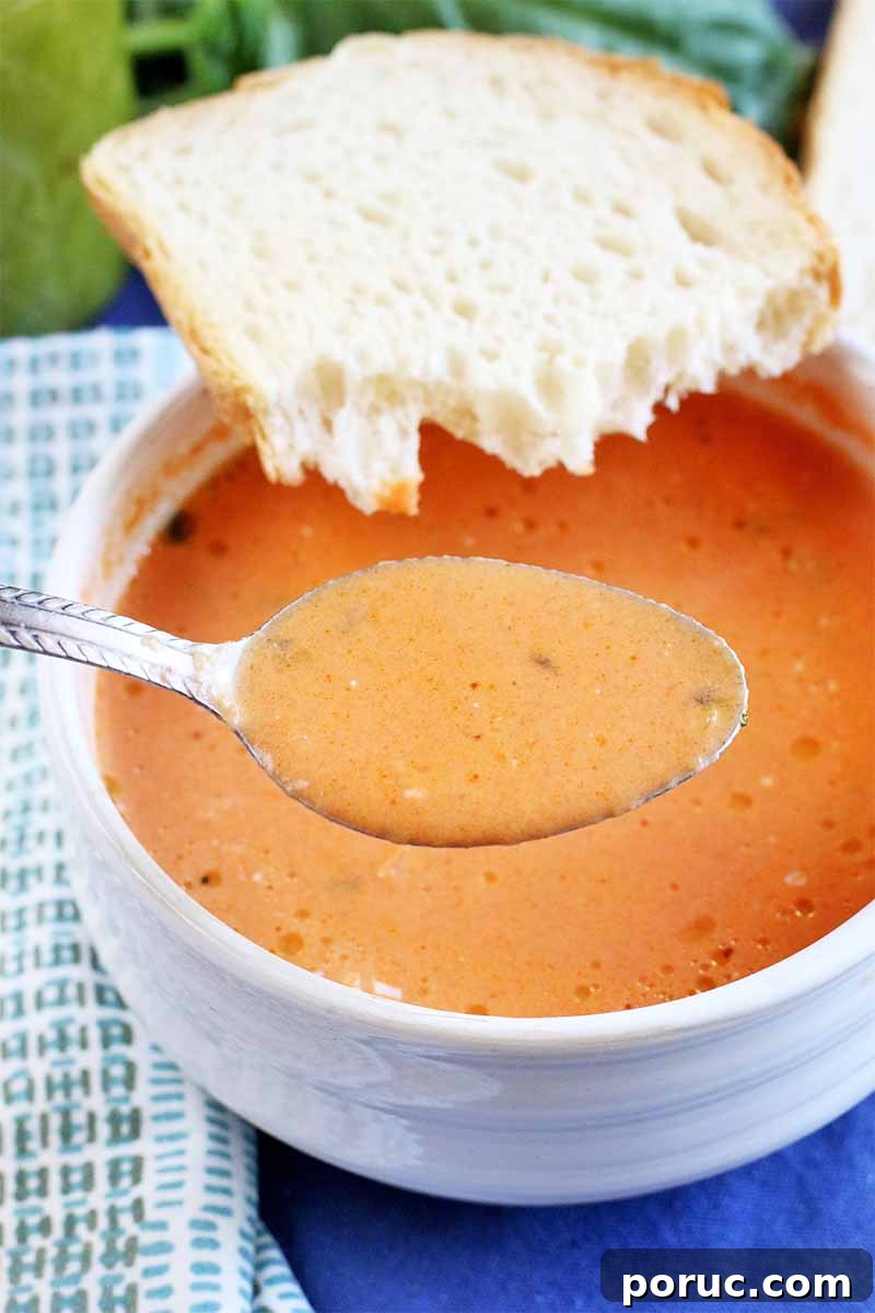 Creamy Tomato Basil Bisque That Elevates Classic Comfort Food 7 A spoonful of pale orange tomato soup is held up to the camera, with a bowl of bisque in the background, with half a piece of sourdough bread balanced on the rim, next to a gray and white patterned cloth napkin, on a blue tablecloth.