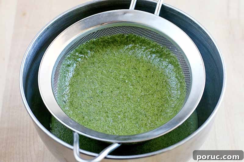 Creamy Tomato Basil Bisque That Elevates Classic Comfort Food 10 A green basil mixture straining into a stainless steel bowl through a stainless mesh strainer, on a beige countertop.