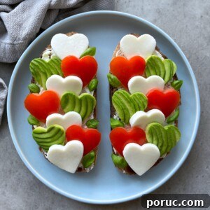 Creamy Avocado & Melted Cheese Toast Recipe 8 Two slices of bread topped with heart-shaped pieces of mozzarella, tomato, and avocado, arranged in a colorful pattern on a blue plate.