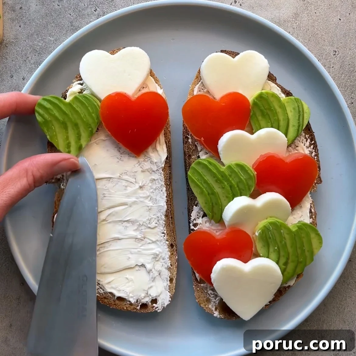 Creamy Avocado & Melted Cheese Toast Recipe 6 Avocado Cheese Toast: Two slices of bread with cream cheese, topped with heart-shaped mozzarella, tomato, and avocado on a plate. A hand is spreading creamy cheese on one slice.