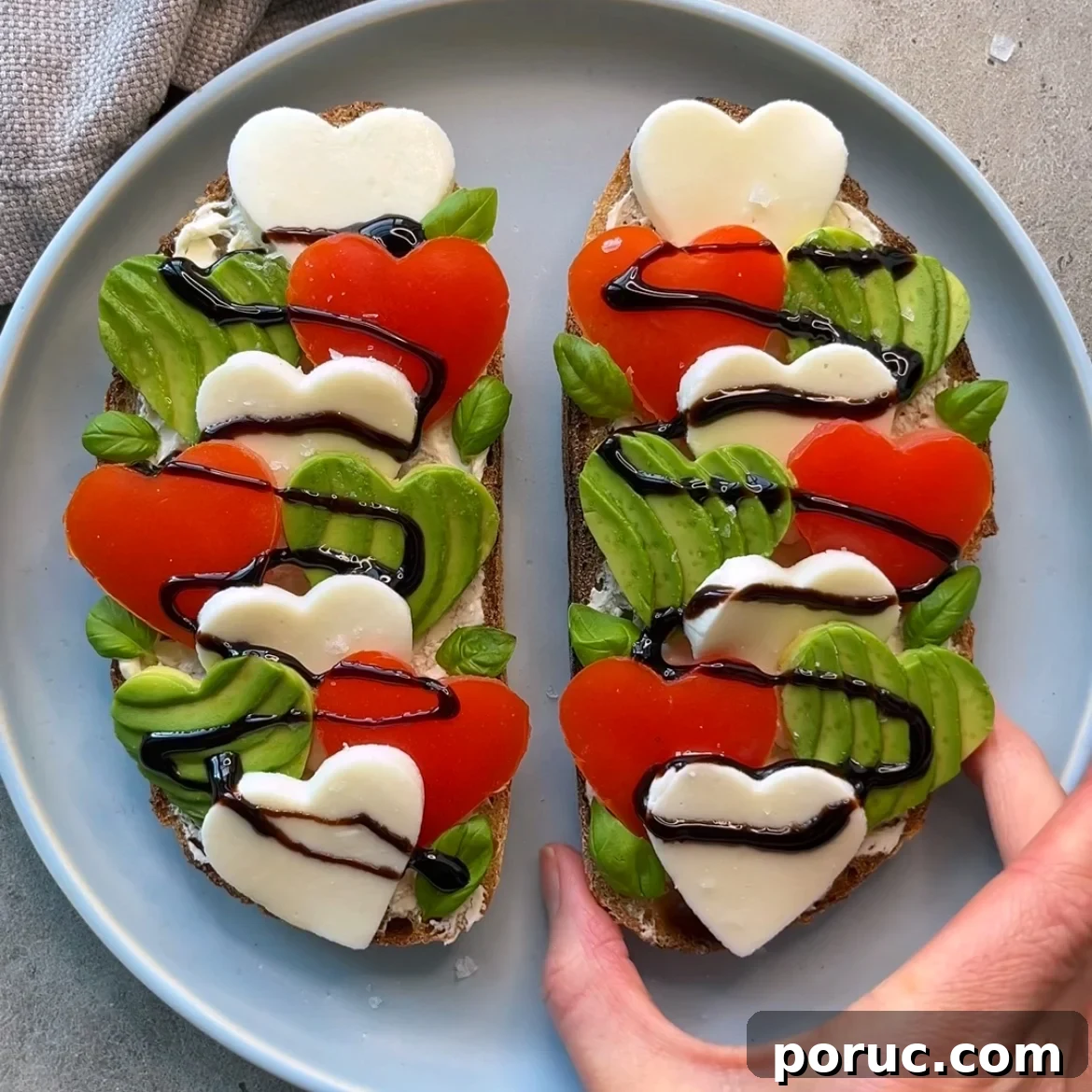 Creamy Avocado & Melted Cheese Toast Recipe 7 Avocado Cheese Toast featuring two slices of bread topped with heart-shaped mozzarella, tomato slices, avocado, basil leaves, and a drizzle of balsamic glaze on a gray plate.