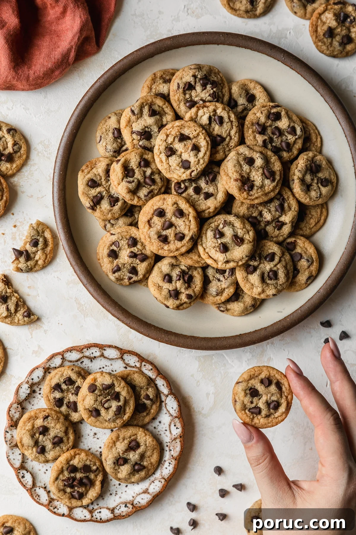Chewy Mini Chocolate Chip Cookie Recipe for Perfect Bites 2 White stoneware plates of mini chocolate chip cookies on a tan counter next to an orange linen, cookies, and a hand holding a cookie.