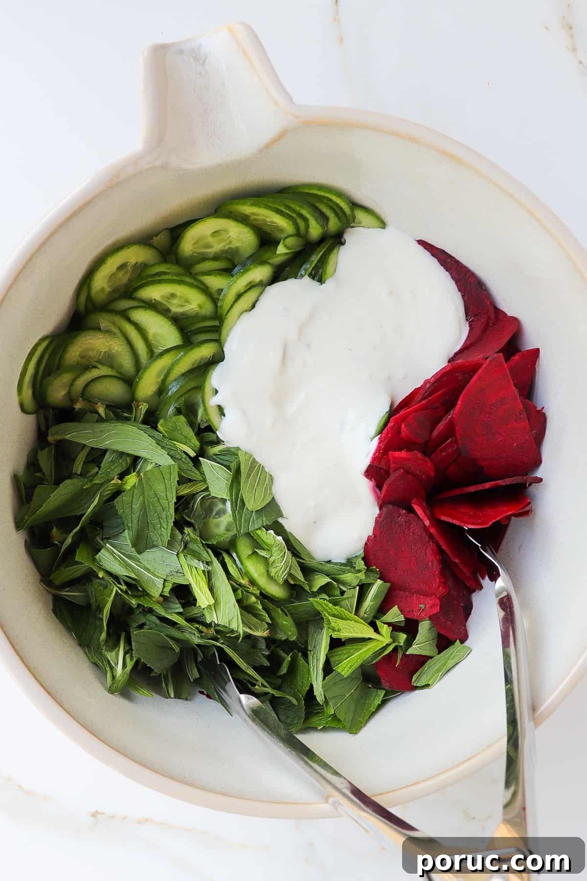 Sliced cucumbers, beetroots, mint leaves and yoghurt dressing in a mixing bowl with metal tongs.