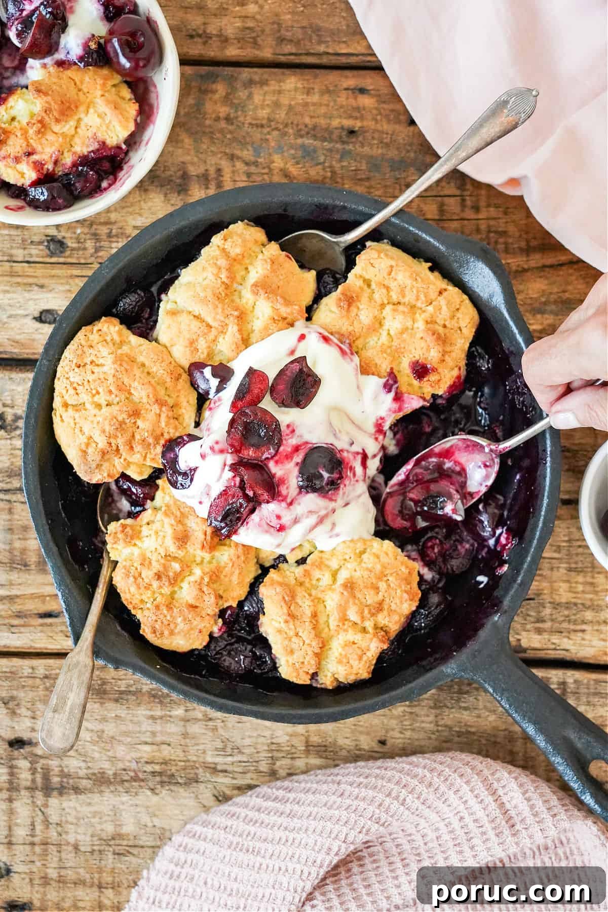 Simple Cherry Cobbler Recipe with Homemade Biscuit Topping 2 Silver spoons in a cast iron skillet with cherry cobbler. Wooden table. Pink cloth.