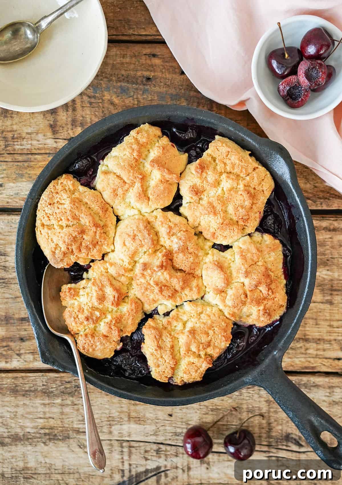 Simple Cherry Cobbler Recipe with Homemade Biscuit Topping 8 Baked cherry cobbler in a cast iron skillet. Wooden surface. White bowl, cherries.