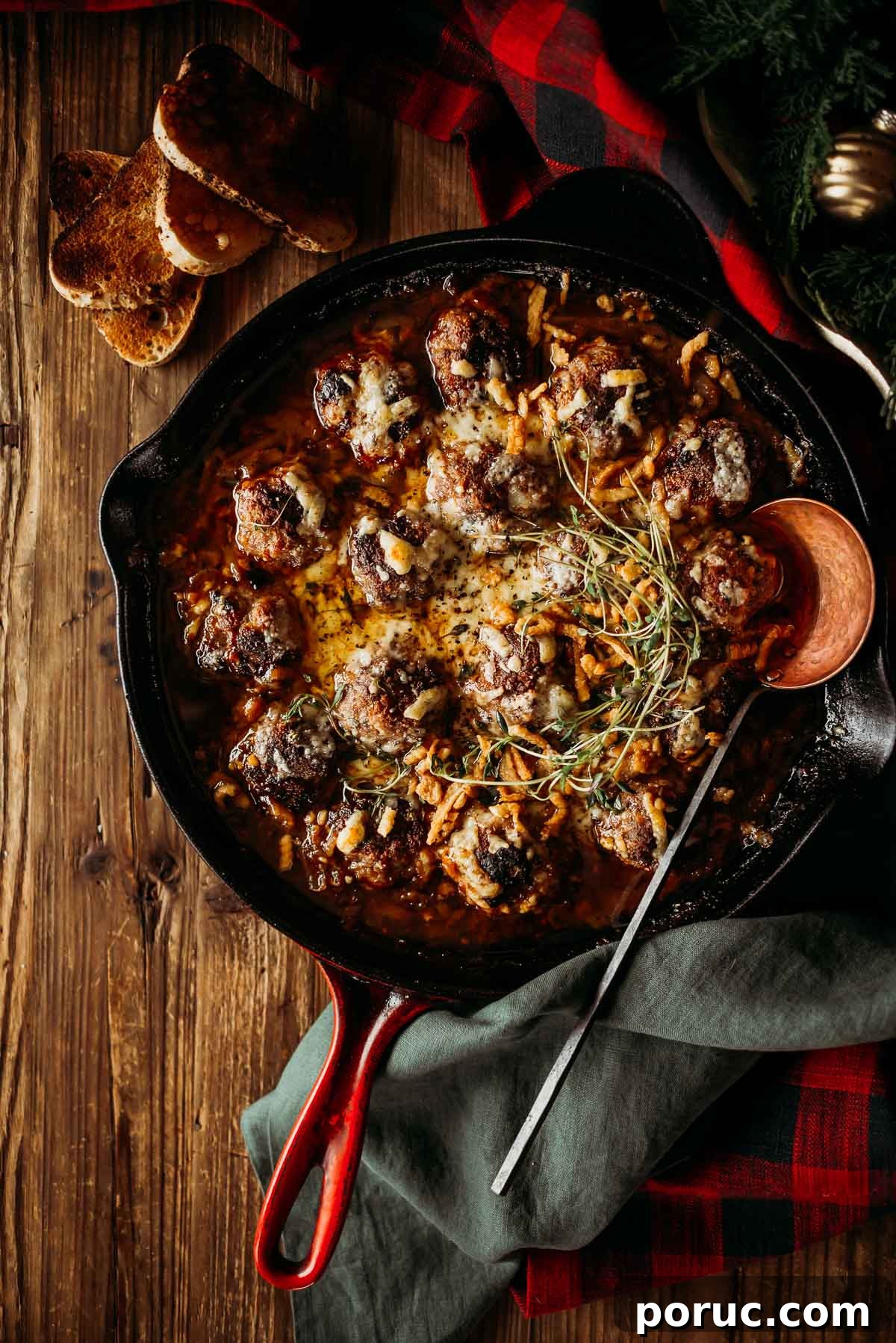 A cast iron skillet with baked french onion meatballs topped with melted cheese and herbs.