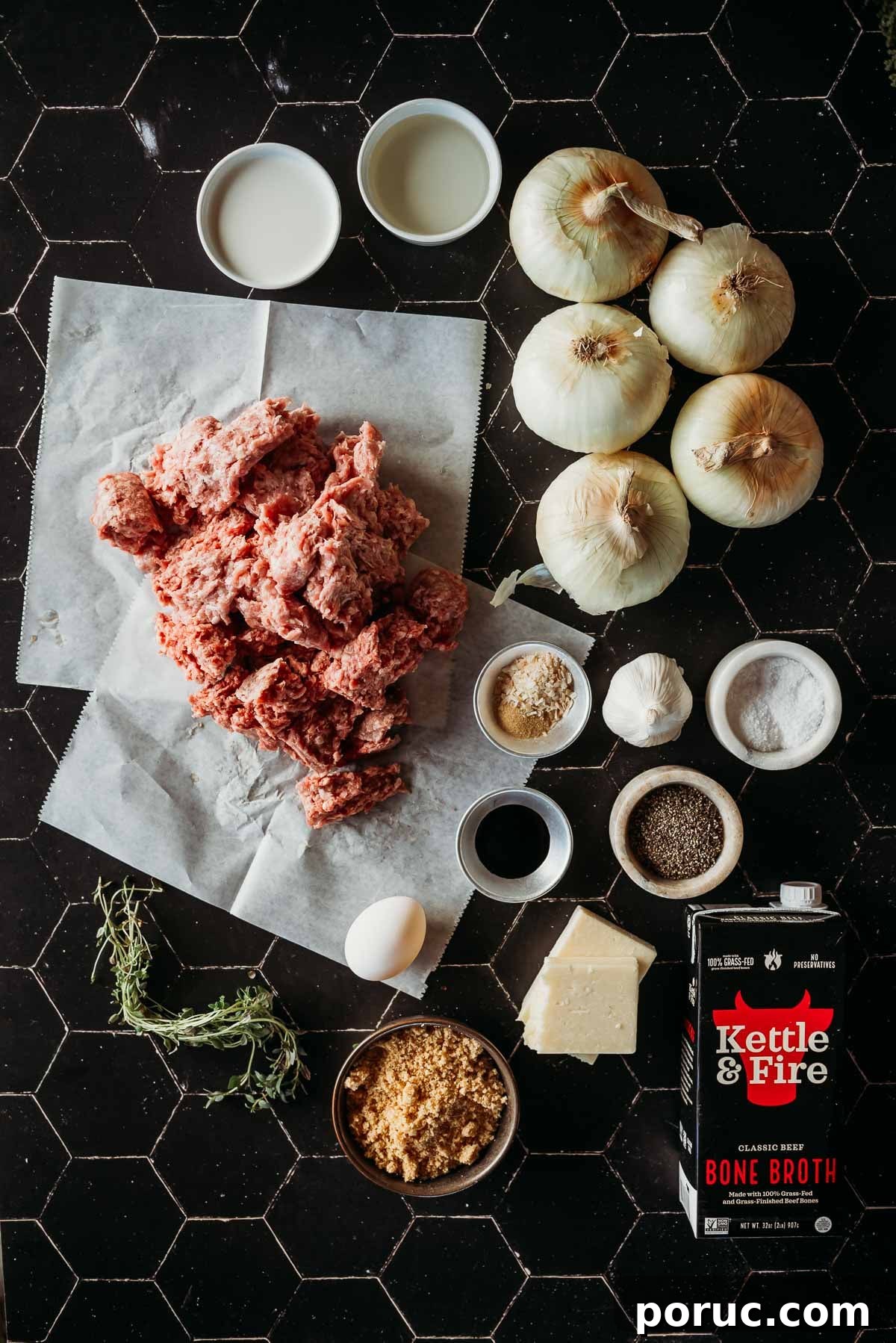 A black tile surface with ground meat, onions, egg, bone broth, herbs, spices, bouillon, and seasonings arranged around.