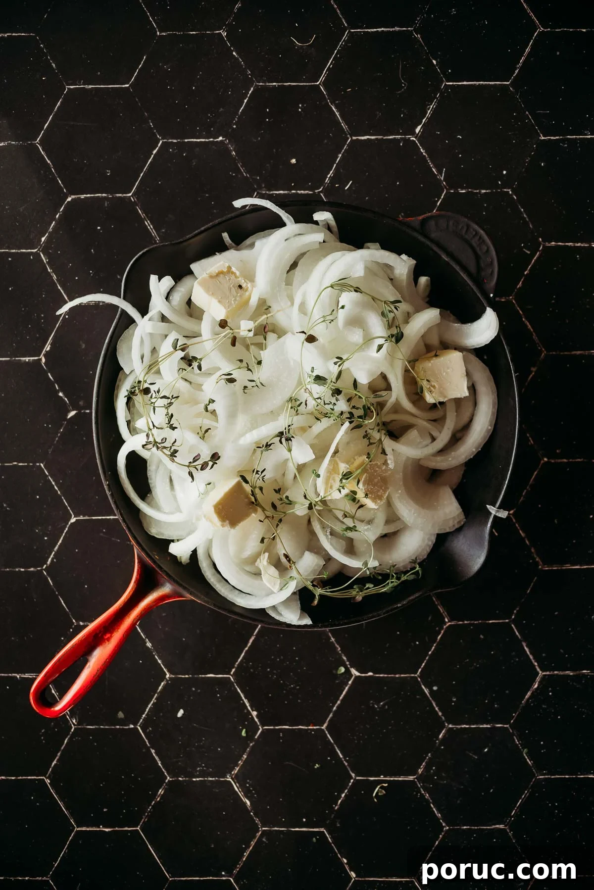 A pan with sliced raw onions, cubes of butter, and sprigs of thyme on a dark hexagonal-tiled surface.