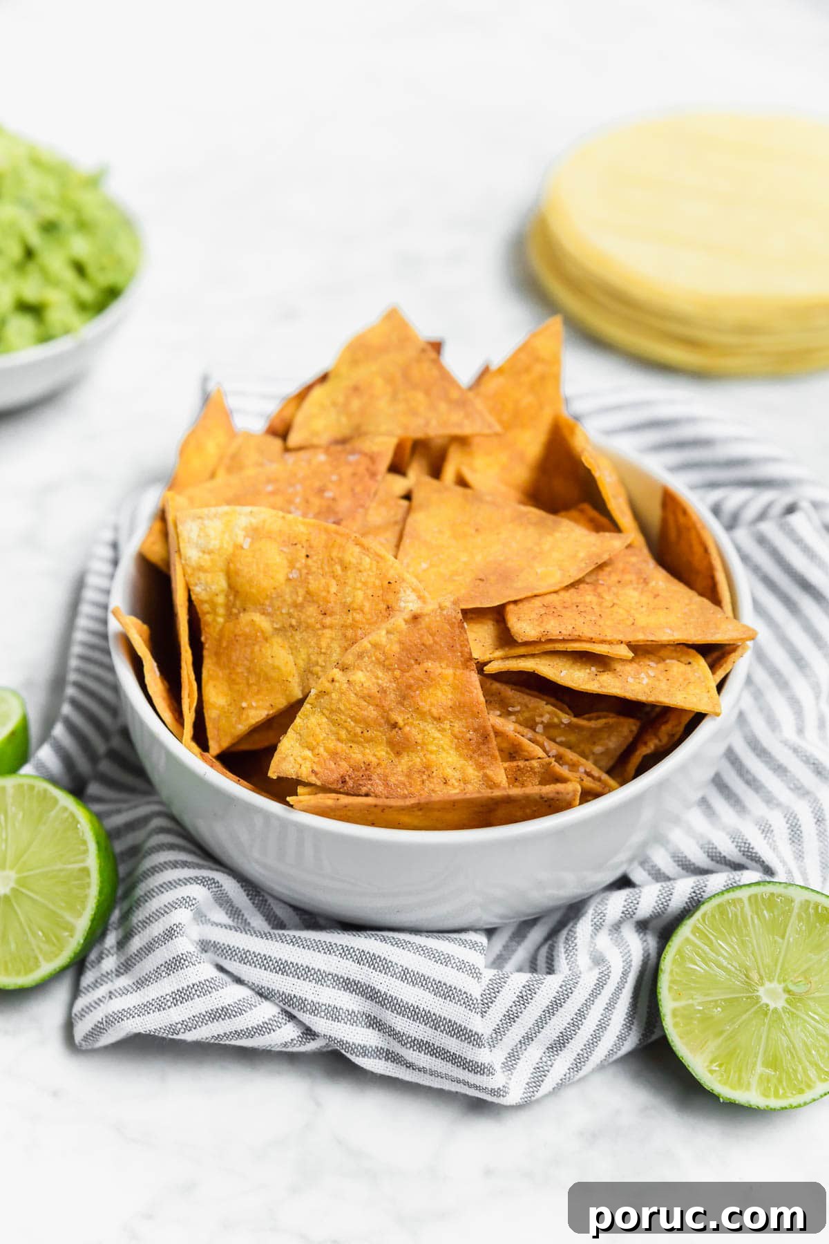 A white bowl filled with golden, crispy Baked Chili Lime Tortilla Chips sits on a striped cloth. Sliced limes and a stack of corn tortillas are nearby, with a bowl of guacamole in the background.