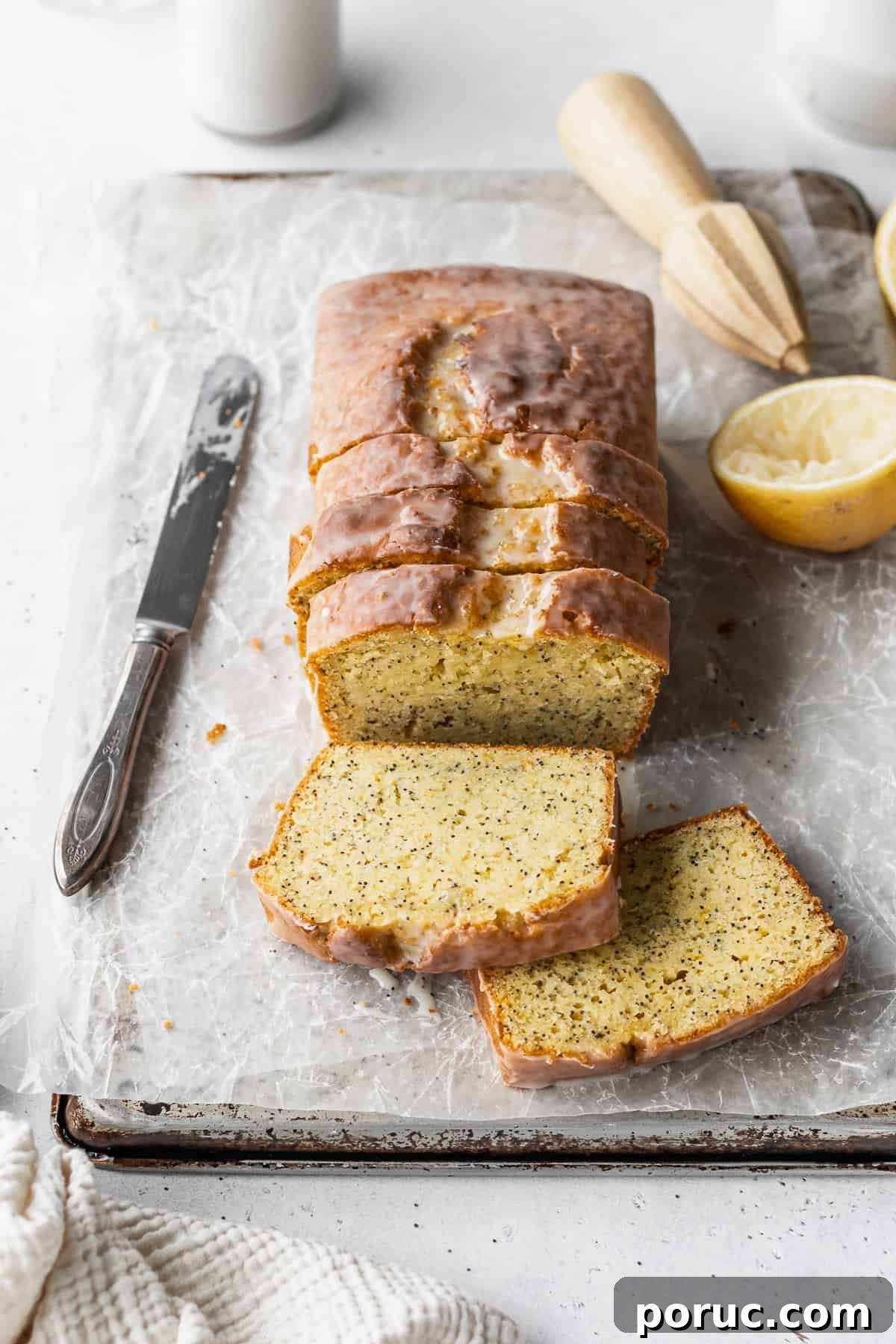 Gluten-Free Lemon Poppy Seed Cake Recipe 2 A sliced loaf of gluten-free lemon poppy seed loaf on a parchment paper lined baking sheet with a knife, half a lemon, and a wooden citrus zester placed around it.