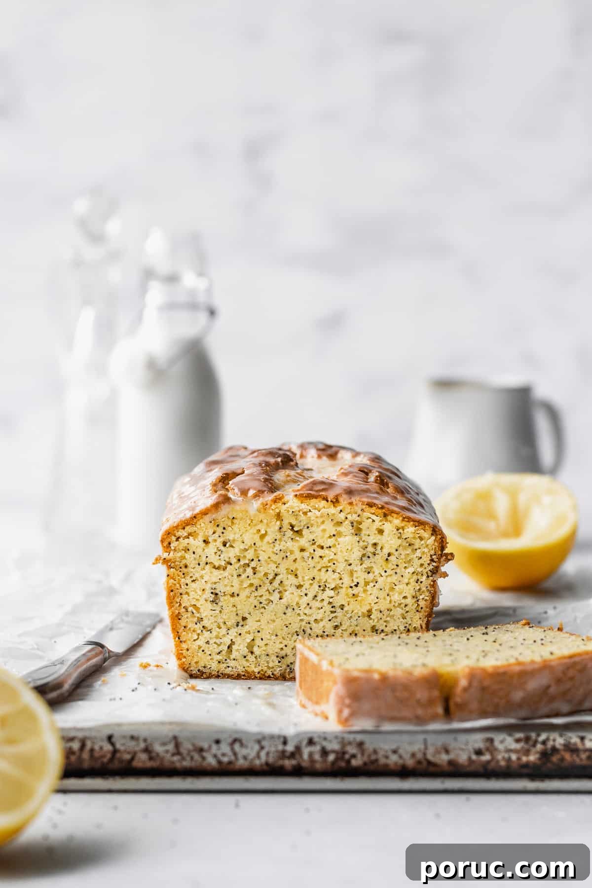 Gluten-Free Lemon Poppy Seed Cake Recipe 3 A sliced loaf of gluten free lemon poppy seed cake on a parchment paper with sliced lemons and milk jugs in the background.