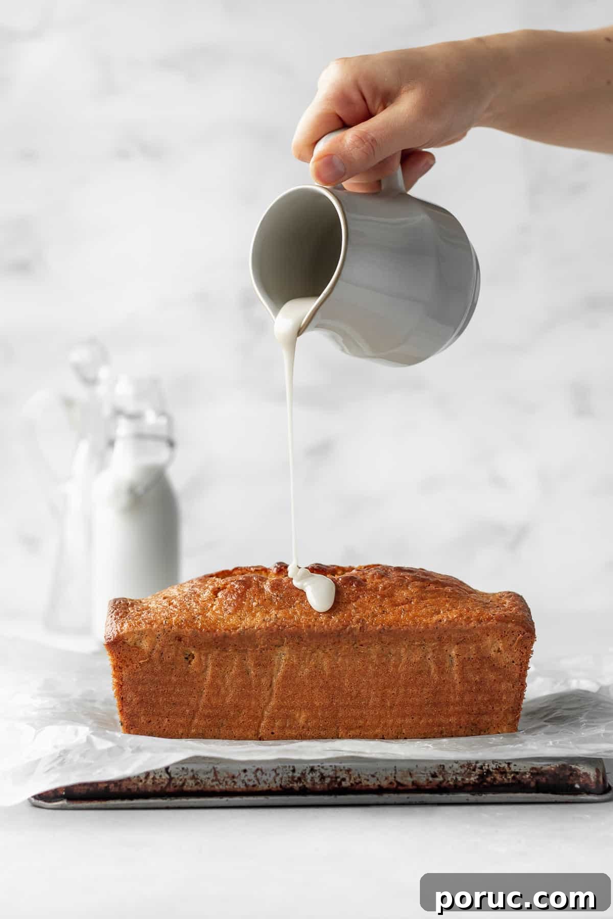 Gluten-Free Lemon Poppy Seed Cake Recipe 4 A hand holding a ceramic jug of lemon icing being poured onto a loaf of gluten free lemon poppy seed cake.