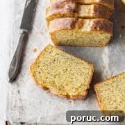 A slice of gluten free lemon poppy seed cake laying on parchment paper with a knife next to it and the sliced loaf behind it.