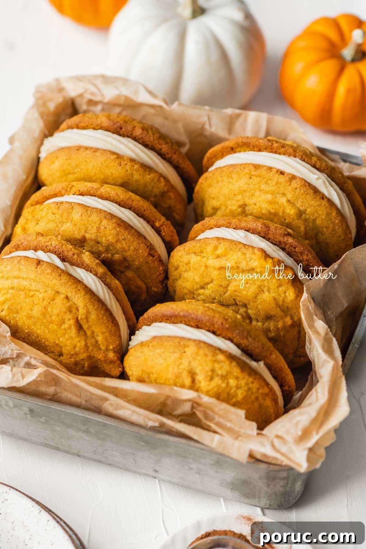 Six pumpkin whoopie pies placed in a brown parchment paper lined metal baking tin with mini orange and white pumpkins placed around it on a white background.