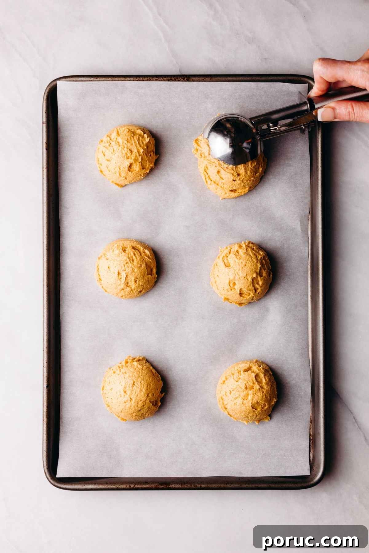 Scooping pumpkin whoopie pie batter onto a parchment paper lined baking sheet on a light grey marbled background.