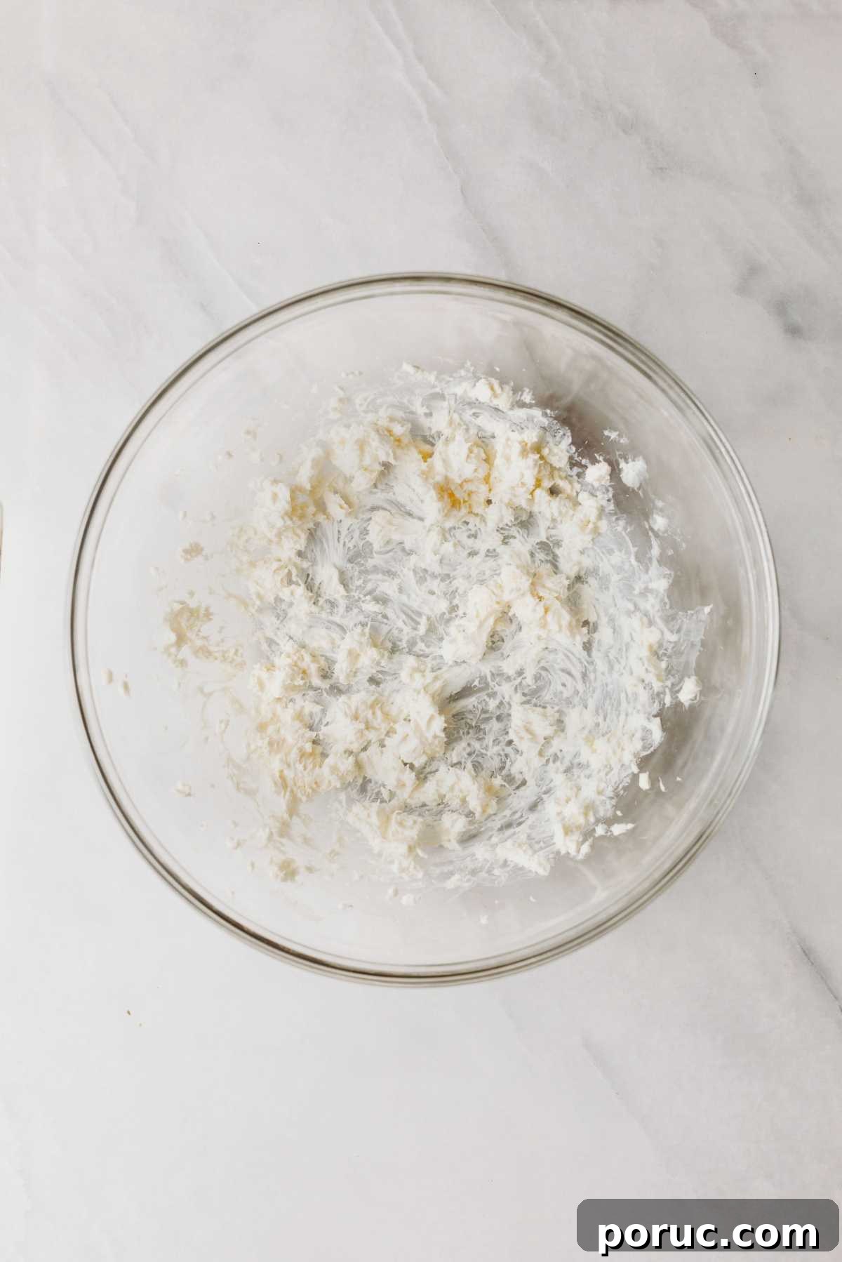 Whisked unsalted butter and cream cheese in a glass mixing bowl on a light grey marbled background.