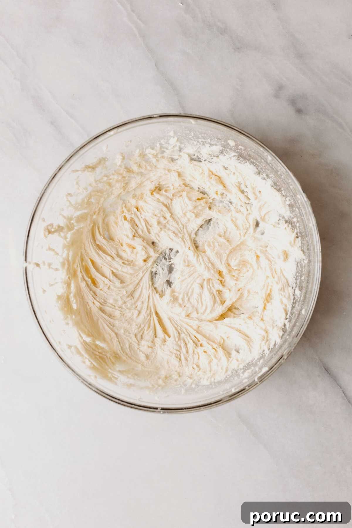 Maple cream cheese frosting in a large glass mixing bowl on a light grey marbled background.