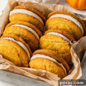 Six pumpkin whoopie pies placed in a brown parchment paper lined metal baking tin with mini orange and white pumpkins placed around it on a white background.