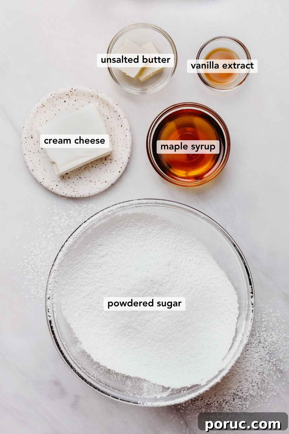 Ingredients in various size bowls labeled as unsalted butter, vanilla extract, cream cheese, maple syrup, and powdered sugar on a white marbled background.