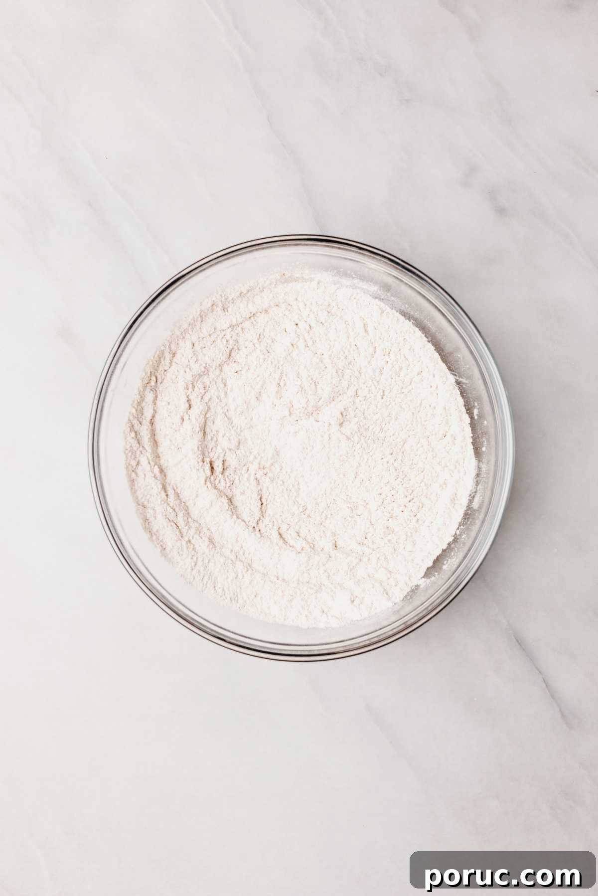 Whisked all purpose flour, baking soda, baking powder, cinnamon, and salt in glass mixing bowl on light grey marbled background.