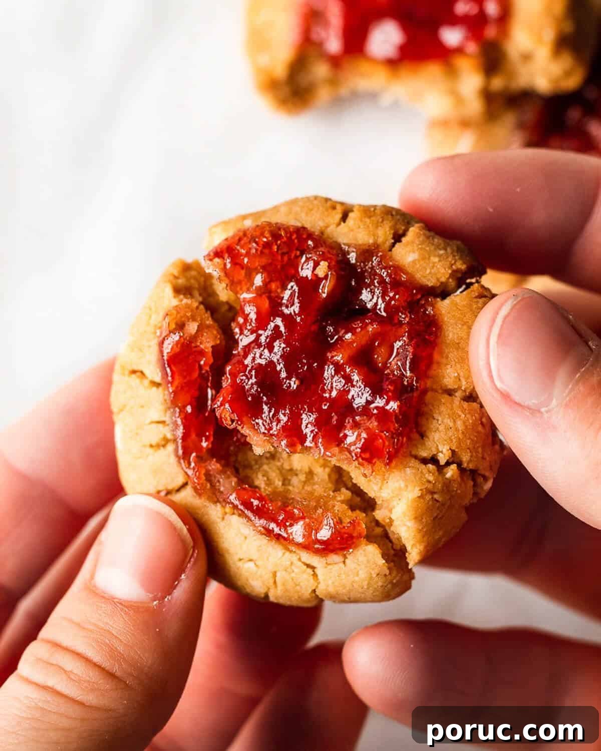 Peanut Butter and Jelly Cookies Recipe — Classic Sandwich Twist 3 Two hands break a jam thumbprint cookie in half with more cookies in the background.